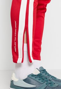 Rode sportbroek met witte zijstrepen en ritsen, met de tekst "REPLAY 'Geen gewone mensen'" afgedrukt op het linkerpantalon.