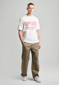 White cotton t-shirt with red text "Manhattan Running Club" and horizontal lines. Beige loose-fitting pants; white sneakers. Simple design.