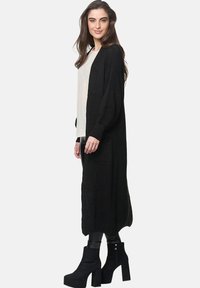 Lång svart stickad cardigan med långa ärmar, sidofickor och ribbad textur, matchad med en ljus skjorta och svarta högklackade stövlar.