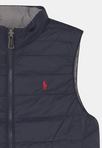 Námořnická puffer vesta s vysokým límcem, plným zipem a vodorovným prošíváním. Na levé straně je vyšívané červené logo.