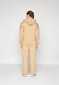 Beige huppari, jossa on leveät hihat ja yhdistelevät collegehousut. Sujuva pinta ja rento istuvuus, esitetty takaa.