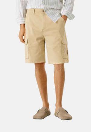 Uomo che indossa pantaloncini cargo beige, una camicia bianca a righe con maniche arrotolate e scarpe marroni slip-on, in piedi con le mani parzialmente nelle tasche.