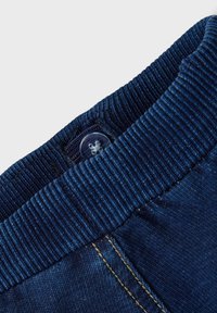 Donkerblauwe denim broek met een geribbelde elastische tailleband en een doorzichtige knop, met zichtbare stikdetails en een zachte textuur.