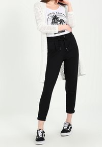 Vit långstickad cardigan, svarta avsmalnande byxor med dragsko och svarta sneakers. T-shirten har grafiskt motiv av palmträd.