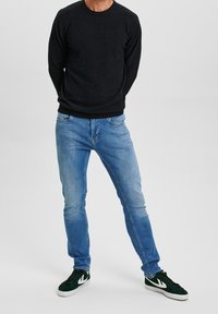 Man med svart tröja, ljusblå jeans och mörkgröna sneakers som står mot en enkel ljus bakgrund.