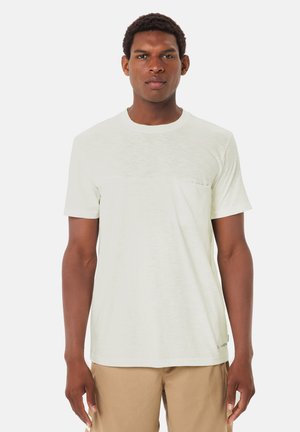 Mann trägt ein schlichtes hellbeiges T-Shirt mit kurzen Ärmeln und beigefarbene Hosen, steht vor einem weißen Hintergrund und schaut nach vorne.