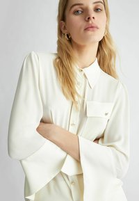 Blusa bianca in tessuto liscio, con colletto, taschino frontale e maniche a campana. Presenta chiusure con bottoni e sottili accenti metallici.