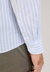Camicia a maniche lunghe a righe azzurro chiaro e bianco, realizzata in un tessuto leggero, con polsini abbottonati e una consistenza sottile.