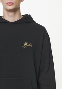 Sudadera con capucha de tejido waffle negro que presenta un bolsillo frontal y un logo bordado en dorado en el lado izquierdo del pecho. Textura suave con un ajuste holgado.