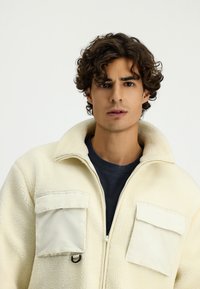 Jasno kremowy polarowy kardigan z zamkiem z przodu, wysokim kołnierzem i dwiema kieszeniami na piersi w jasnym materiale. Teksturowana powierzchnia o swobodnym designie.