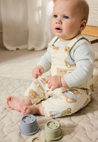 Salopette pour bébé en coton avec un imprimé d'ours en beige et bleu, accompagnée d'un t-shirt à manches longues rayé bleu ; trois gobelets empilables colorés à proximité.