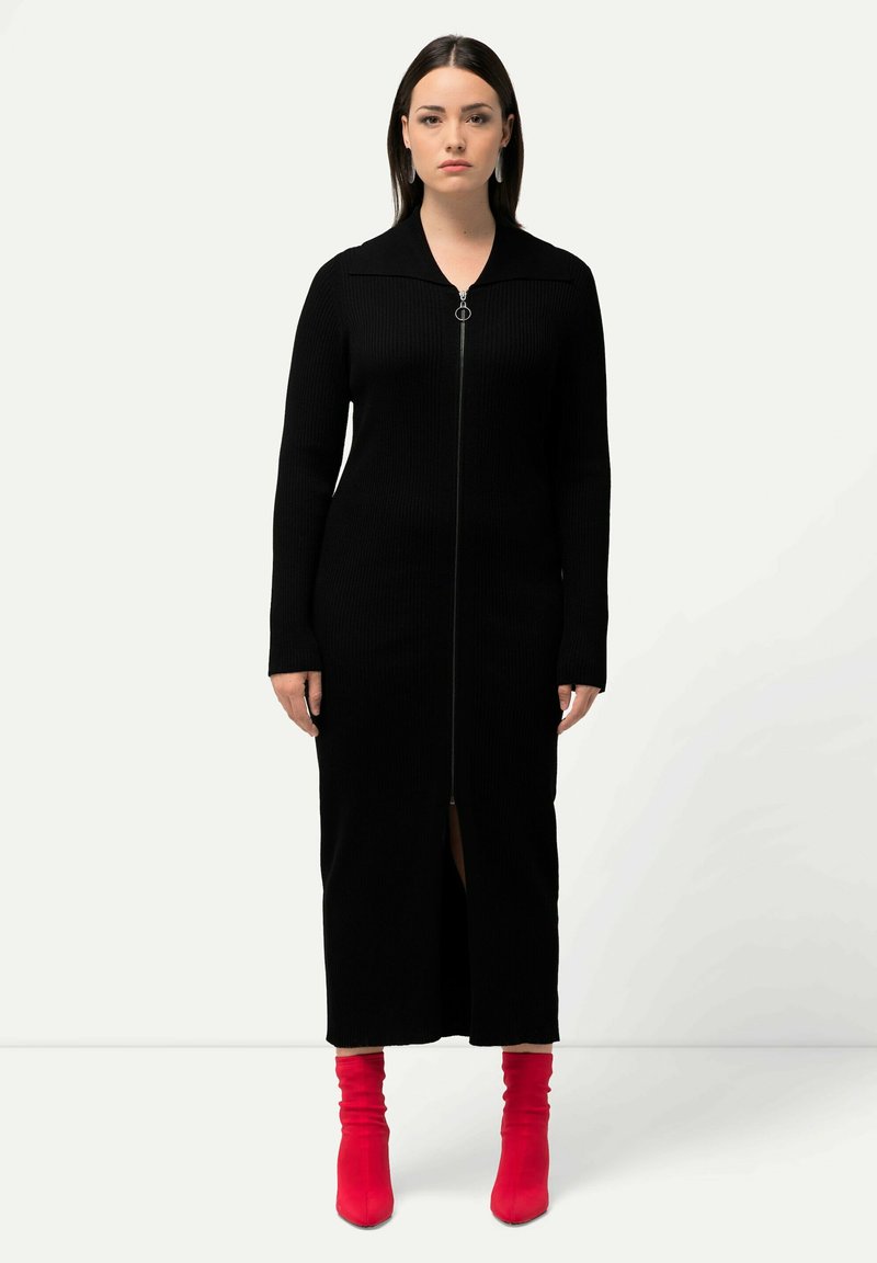 Robe en maille côtelée noire avec fermeture éclair, manches longues et fente à l'avant. Associée à des bottes à talons larges rouges.