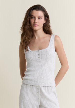 Mujer con cabello largo y ondulado de color castaño, vistiendo un top blanco sin mangas de punto con botones y pantalones holgados a juego, frente a un fondo beige liso.