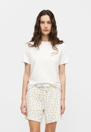 Vrouw met lang krullend haar, die een wit shirt met korte mouwen en witte korte broek met kleine gele citroenmotieven draagt, staand tegen een effen achtergrond.