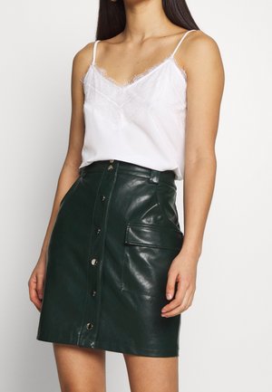 Femme portant un caraco blanc bordé de dentelle et une jupe en cuir vert foncé boutonnée sur le devant avec une grande poche latérale.
