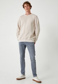 Sudadera color beige claro con cuello redondo, con puños y dobladillo acanalados. Combinada con jeans azul claro y zapatillas blancas con suelas marrones.