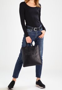 Schwarze Leder-Shopper-Tasche mit geprägt "DKNY" Logo, quadratischer Form, zwei kurzen Tragegriffen, glatter Textur. Getragen zu lässigen Jeans und schwarzen Schuhen.