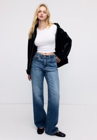 Femme blonde portant une veste en cuir noire sur un crop top blanc, un jean large bleu, et des ballerines noires, se tenant devant un fond blanc.
