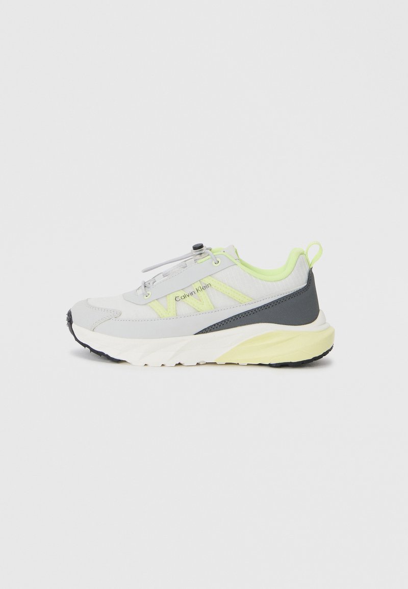 Chaussure de sport grise avec des accents jaune fluo, tige en maille et semelle texturée. Présente un design épuré et une languette de tirage au niveau du talon.