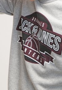 Sudadera gris con un gráfico central que presenta el texto "Jack & Jones", un diseño de baloncesto y una pancarta que dice "Originales".