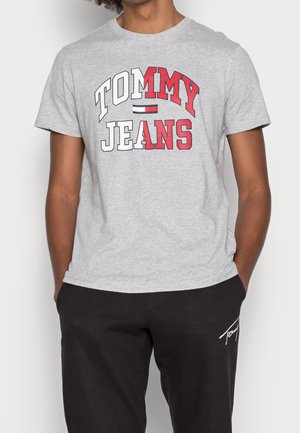Henkilö pukeutunut vaaleanharmaaseen "Tommy Jeans" t-paitaan, jossa on punaiset ja valkoiset kirjainmerkit, sekä mustiin housuihin, joissa on pieni valkoinen logo vasemmassa reidessä.