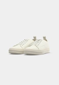 Låga off-white sneakers med snören, gummisula och dragremmar, visade sida vid sida på en ren vit bakgrund.