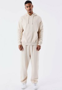 Beige hoodie en bijpassende joggingbroek gemaakt van zacht, gestructureerd materiaal. Heeft een kangoeroezak en geribbelde manchetten, gecombineerd met witte sneakers.