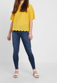 Gul broderad blus med vågig nederkant, matchad med mörkblå tajta jeans och vita sandaletter på en enfärgad bakgrund.