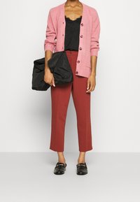 Cardigan rosa testurizzato con bottoni, top in pizzo nero, pantaloni cropped color ruggine e mocassini neri lucidi. Tiene in mano una borsa nera stropicciata.