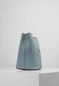 Zachte blauwe leren emmer tas met een gestructureerde afwerking, voorzien van een gouden hardware accent en een open bovenkant, zittend op een witte sokkel.