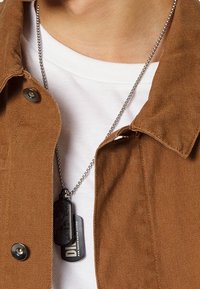 Brown fabric jacket with button closure, over a white shirt. A silver-chain necklace shows two dog tags, one matte black and one with text.