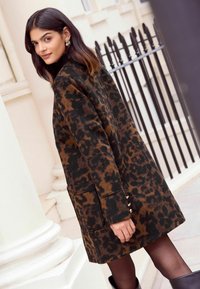 Ogrtač s leopard uzorkom u smešnoj i crnoj boji, s bočnim džepovima i naglascima na dugmadima. Mekana tekstura i ležeran kroj. Model gleda bočno.
