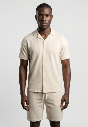Homme debout portant une chemise beige à manches courtes boutonnée et un short assorti avec cordon de serrage sur un fond uni clair.