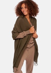 Châle vert olive avec des franges, fabriqué en tissu doux, porté sur une tenue brun clair, avec un design à boutons et une coupe décontractée.