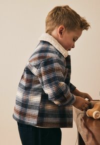 Karo jaka zila, brūna un krēmkrāsas toņos ar balto šerpa apkakli. Kokvilnas audums, brīvā piegriezumā un redzama tekstūra.