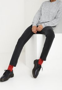 Grå stickad tröja, svarta slim-fit jeans och svarta snörskor med gula sömmar. Röda strumpor synliga ovanför skotopparna. Enkel bakgrund.