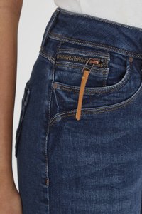 Jeans de mezclilla azul oscuro con una cintura alta, que presentan un bolsillo con cremallera con una lengüeta de cuero marrón y costuras naranjas contrastantes.