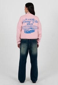 Femeie văzută din spate, purtând un pulover roz cu text albastru „In a Rush Racing Club Berlin” și o grafică cu mașină, asortat cu blugi largi închiși la culoare.