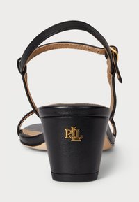 Must ette sandaalid kitsaste kontidega, nahkrihmadega ja kuldse logoga taga. Heledatäpiline esmärk, millel on sile tekstuur.