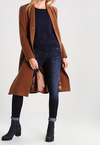 Abrigo largo marrón de lana con dos botones, camiseta azul marino, jeans oscuros y botas negras hasta el tobillo. Textura suave con un interior forrado de satén.