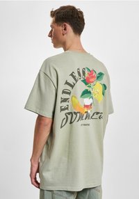 Camiseta holgada verde claro con un estampado gráfico de flores y frutas, con las palabras "VERANO INFINITO" estilizadas debajo.