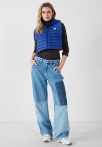 Chaleco acolchado azul con detalles rojos, cremallera frontal, usado sobre un jersey de cuello alto negro, combinado con vaqueros de pierna ancha de parches azul claro y zapatillas blancas.