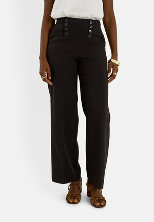 Mujer con pantalones negros de cintura alta con botones decorativos, blusa blanca metida por dentro, sandalias de tacón con estampado de leopardo y joyas doradas en las manos.