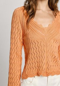 Oranje gebreide trui met een zigzagpatroon, lange mouwen, een geschulpte halslijn en een getextureerde afwerking. Draagt lichtgekleurde broek.