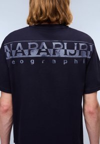 Mørkeblå t-shirt med stort gråt tekst "NAPAPIJRI" og "geography" på ryggen, korte ærmer og en blød bomuldsstof tekstur.