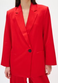 Rot taillierter Blazer mit Reverskragen, ein schwarzer Knopfverschluss, zwei seitliche Taschen und leicht übergroßen langen Ärmeln. Glatte Stofftextur.