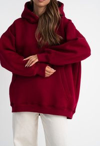 Felpa oversize bordeaux realizzata in morbido misto cotone, con tasca frontale, cappuccio con coulisse e polsini a coste. Abbinata a pantaloni chiari.