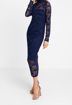 Robe midi en dentelle bleu marine avec manches longues et col montant, associée à des sandales à talons blocs nude avec bride à la cheville.