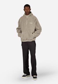 Szürke bolyhos kapucnis pulóver félig cipzárral, elülső zsebbel és reflektív logóval, fekete nadrággal és fekete, fehér talpú sportcipőkkel párosítva.