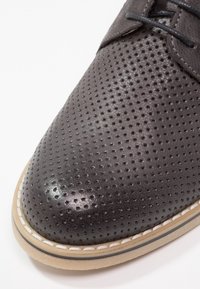 Scarpa da uomo in pelle perforata di colore grigio scuro con texture liscia, punta rotonda, lacci neri e suola in gomma beige a contrasto con dettagli di cucitura.